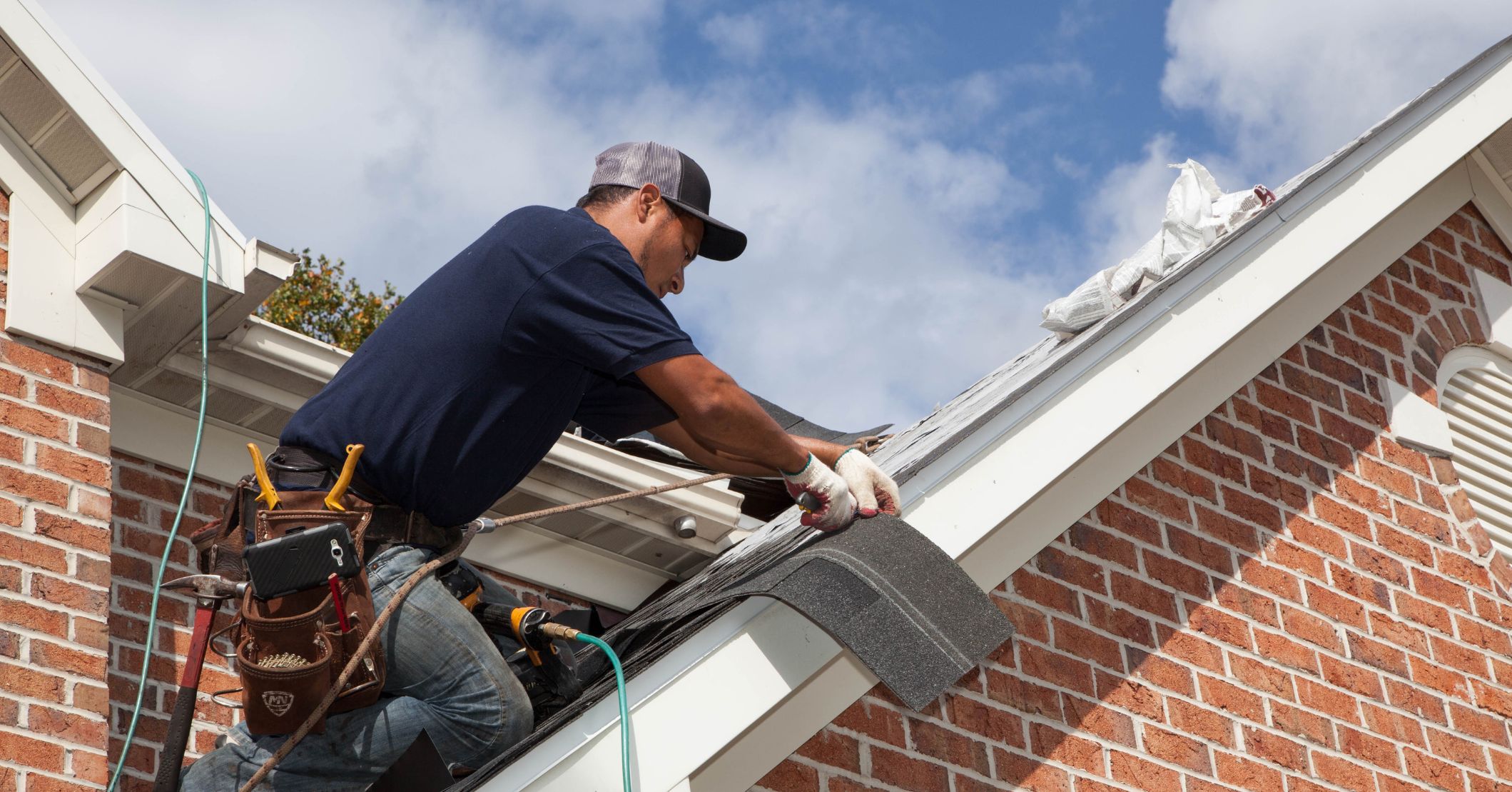 A roofing professional wearing a tool belt and safety gear carefully installing shingles on a brick house.