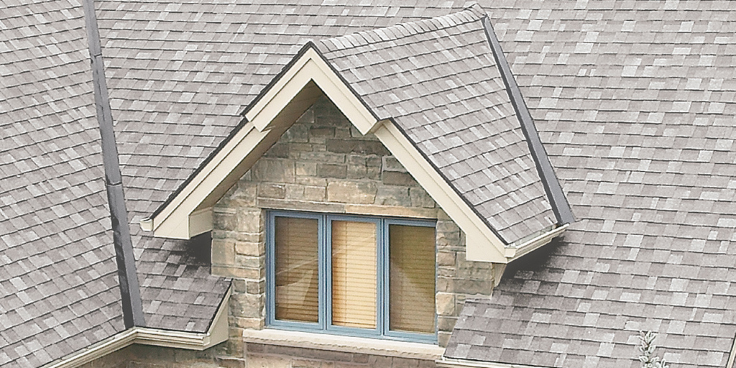 A recessed dormer set into a gray-shingled roof, featuring a triangular gable and stone siding around double windows."