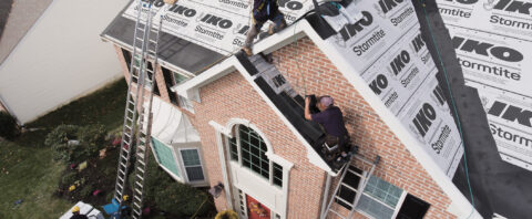 Roofers installing underlayment and drip edge on a two-story brick home roof using ladders and safety harnesses.