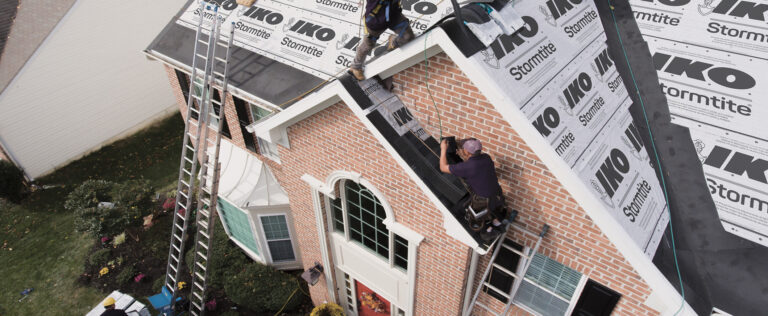 Roofers installing underlayment and drip edge on a two-story brick home roof using ladders and safety harnesses.