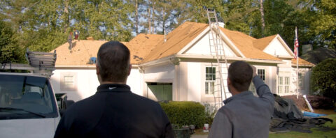 Two men, seen from behind, stand in front of a house that is undergoing a roof replacement; one man is pointing up towards the roof where a ladder rests and a roofer is working.