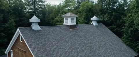 An aerial view of a dark-shingled, barn-style roof featuring a central cupola and two pointed side vents, set against a backdrop of a dense green forest.