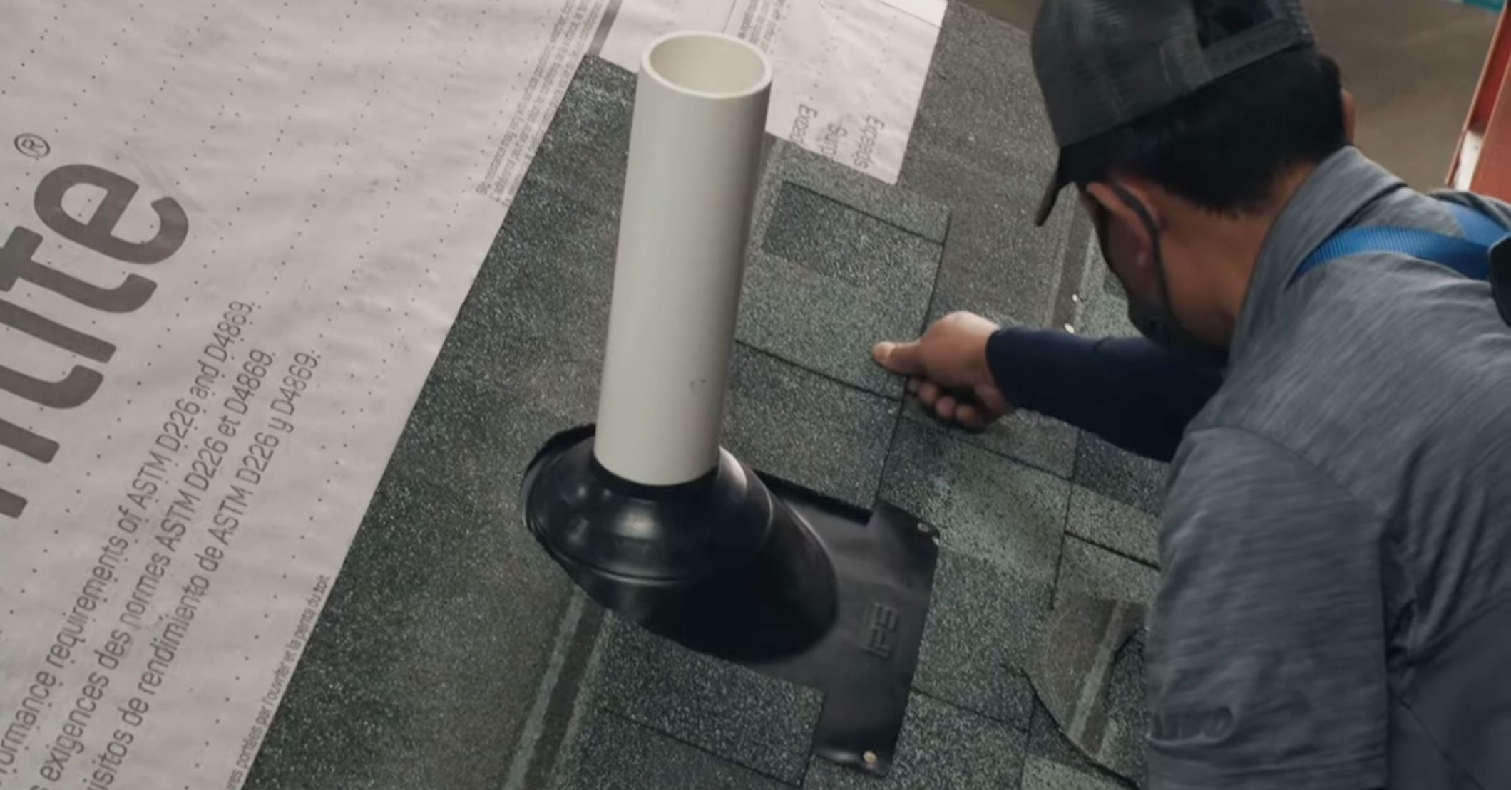 A roofer is carefully installing shingles on a roof around the base of a black plumbing vent boot that surrounds a white pipe.