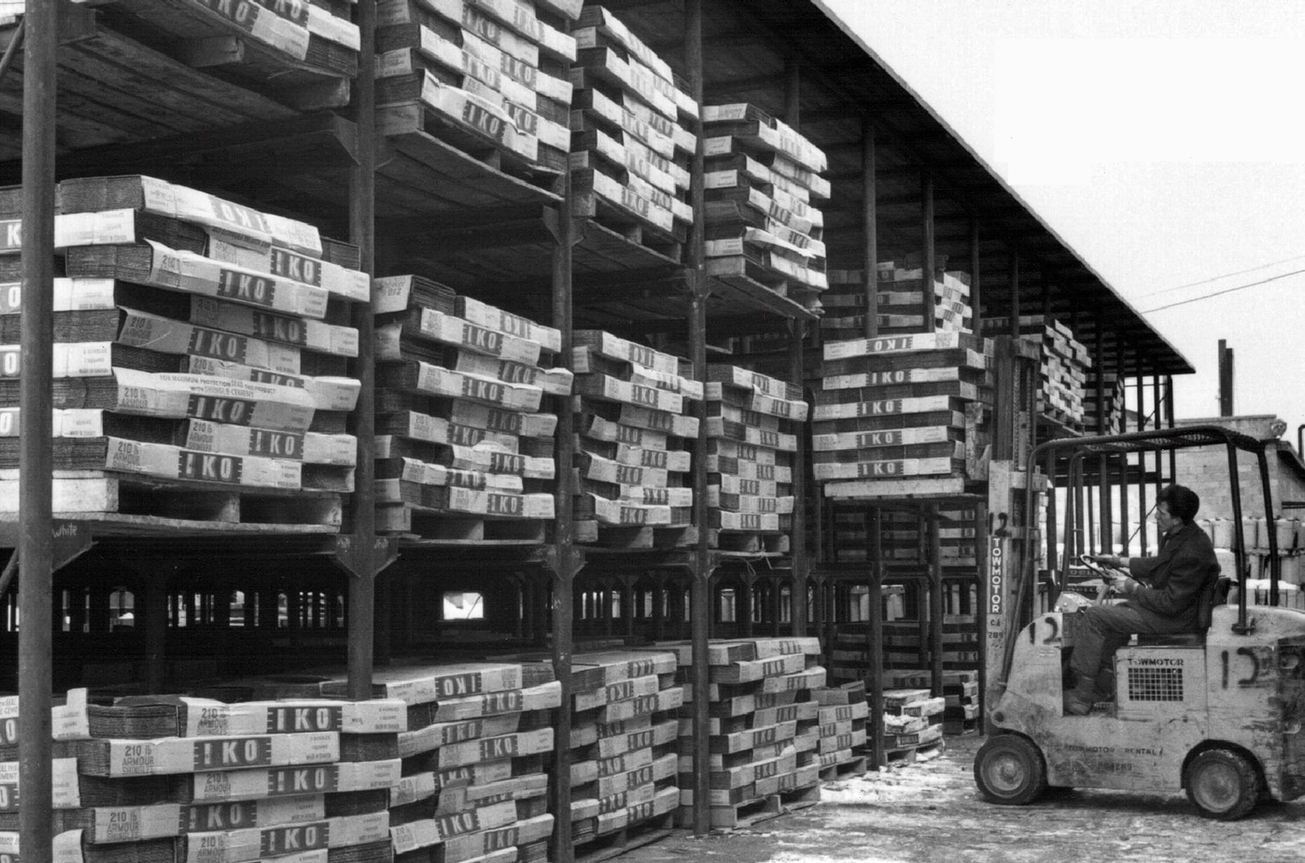 Black-and-white archival warehouse photo showing stacked IKO shingle bundles on racks and a forklift operator moving pallets