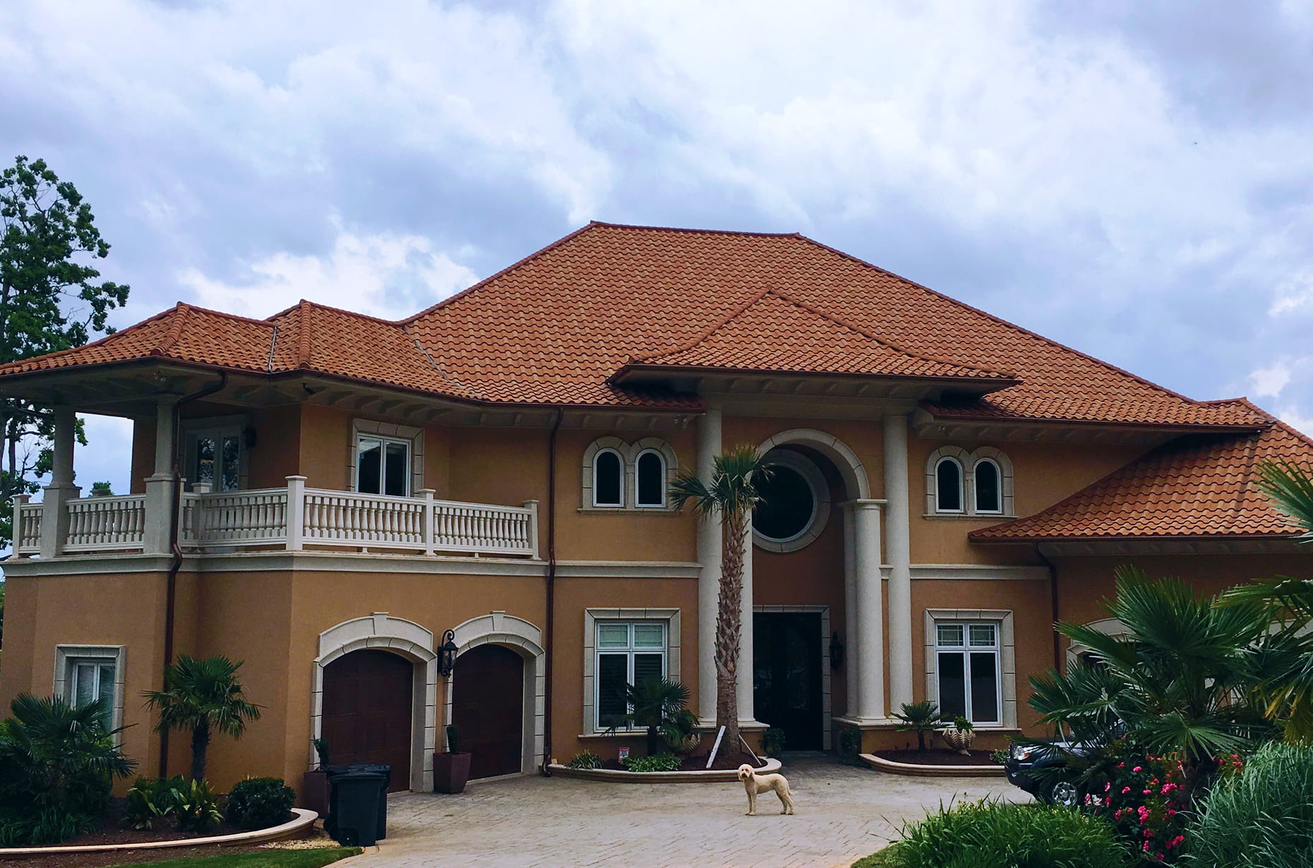 Mediterranean-style stucco mansion with terracotta tile-style DECRA metal roof and small dog standing on driveway