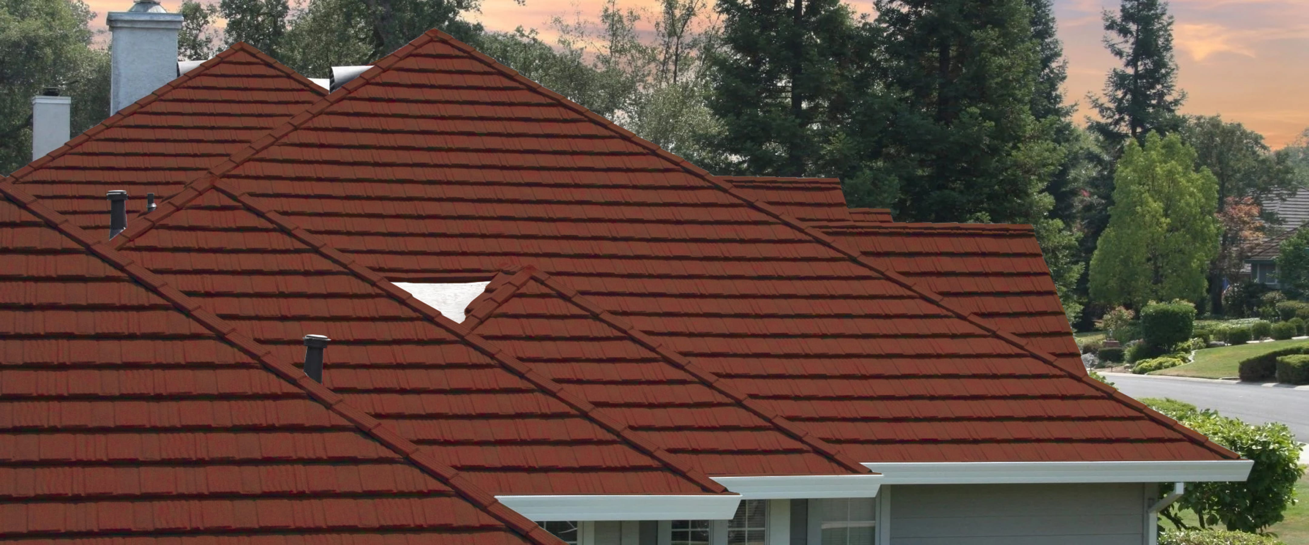 Rust-red metal shingle roof on a house with several gables, with tall trees and a sunset sky behind