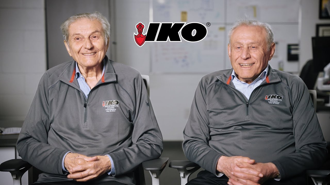 Two smiling older men in gray IKO quarter-zip jackets sitting side by side, with the IKO logo above them