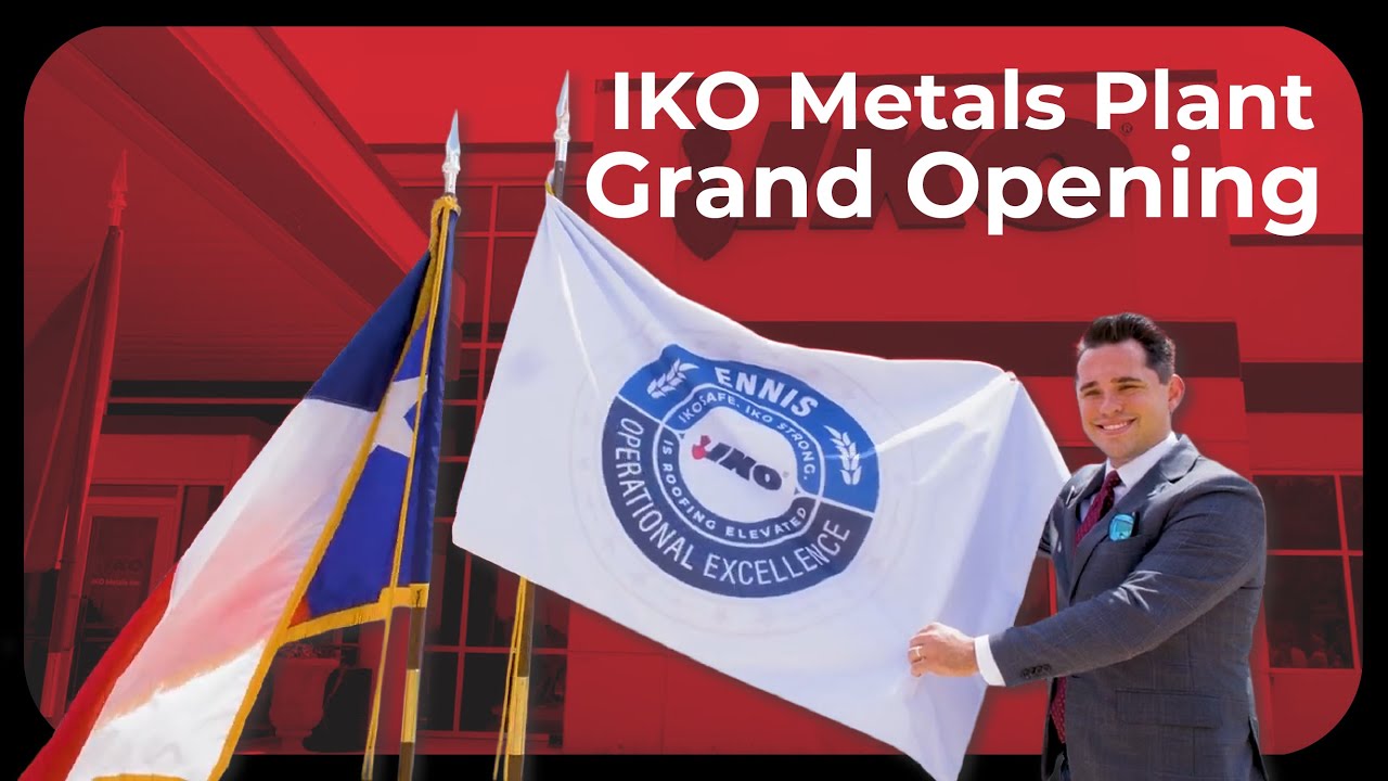 IKO Metals Plant Grand Opening: a smiling man in a suit holding a white Ennis Operational Excellence flag next to a Texas flag