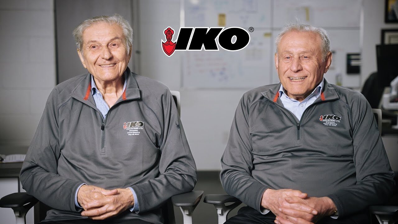 Two smiling older men in gray IKO quarter-zip jackets sitting side by side, with the IKO logo above them