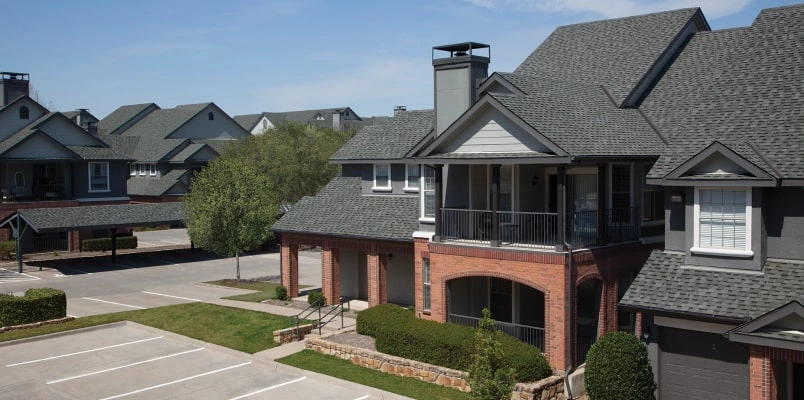 A charming two-story residential building in Cambridge, featuring cool colors with red brick and gray siding. It boasts a balcony and chimney, plus it's adjacent to a parking area surrounded by neatly trimmed bushes and trees.