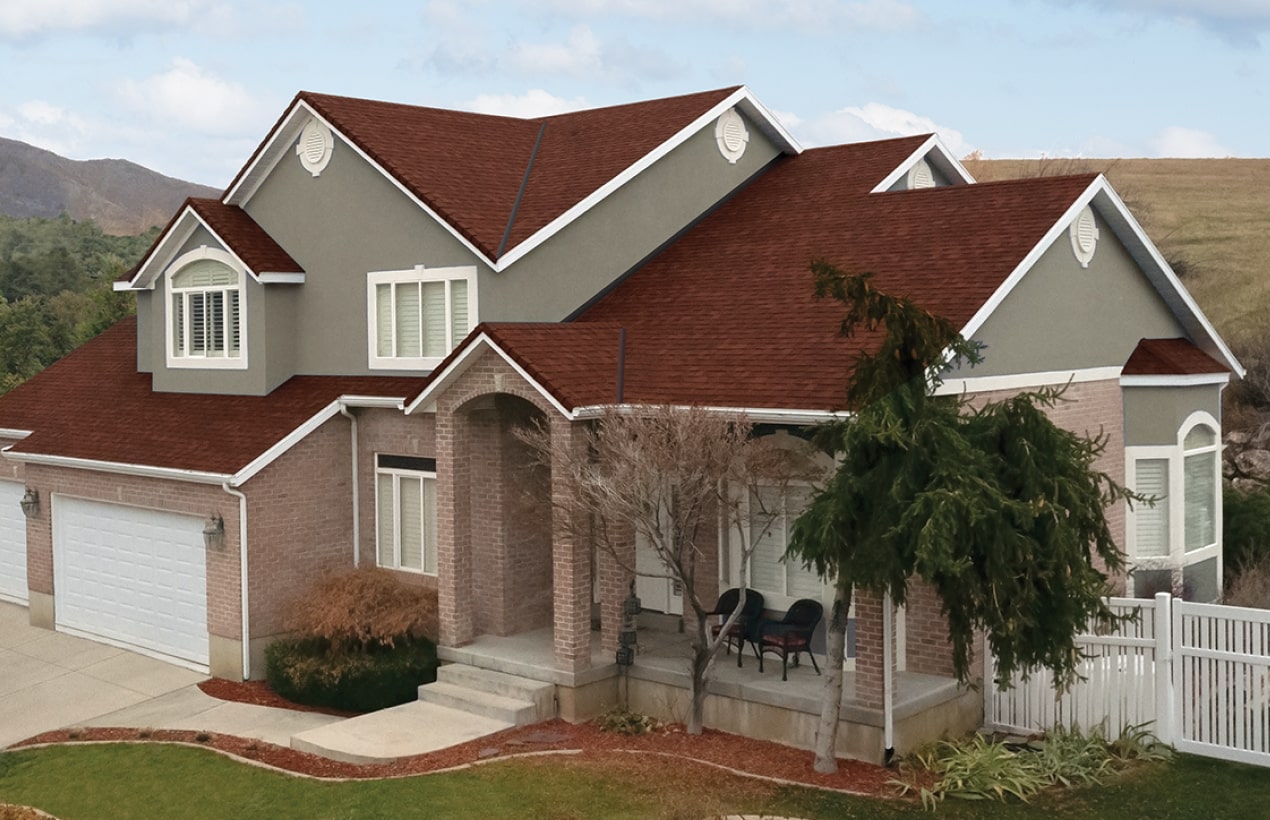 This two-story house boasts a distinctive Monaco Red and gray brick exterior, complemented by its red roof and two-car garage. The front porch, perfect for leisurely afternoons, features two chairs, while a small tree adds charm to the yard.