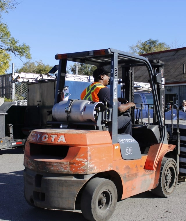 A person wearing a reflective vest operates an orange forklift outdoors, surrounded by vehicles and buildings under a clear blue sky, efficiently maneuvering with the precision warranted in such bustling environments.