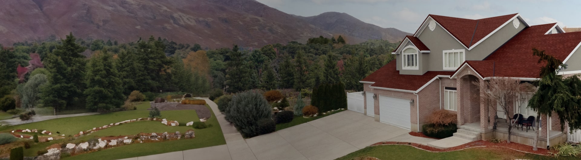 Casa de techo rojo y exterior gris, que inspira en cada aspecto de su diseño, rodeada de un jardín bien cuidado con entrada para autos y montañas con vegetación exuberante al fondo.