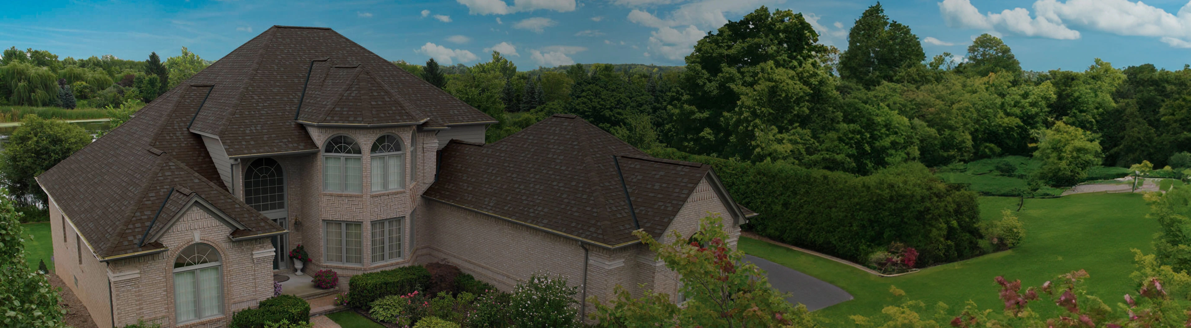 Large brick house with a dark roof, surrounded by lush green trees and bushes under a partly cloudy sky, reminiscent of an architectural style board. A driveway is visible on the right.