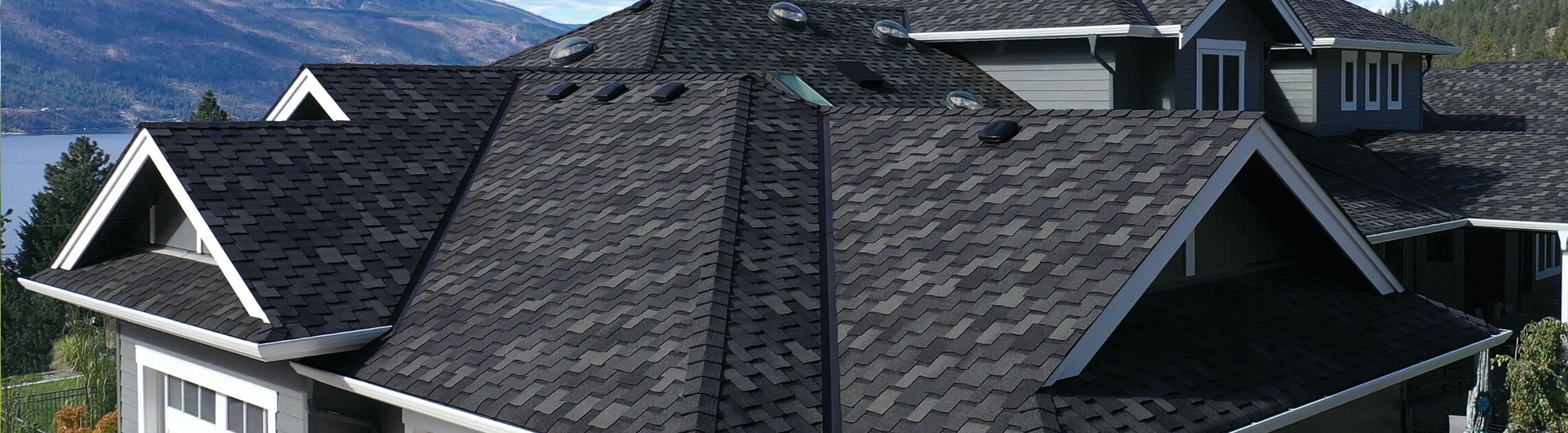 Techo de tejas gris oscuro con tragaluces en una casa junto a un lago, con montañas al fondo