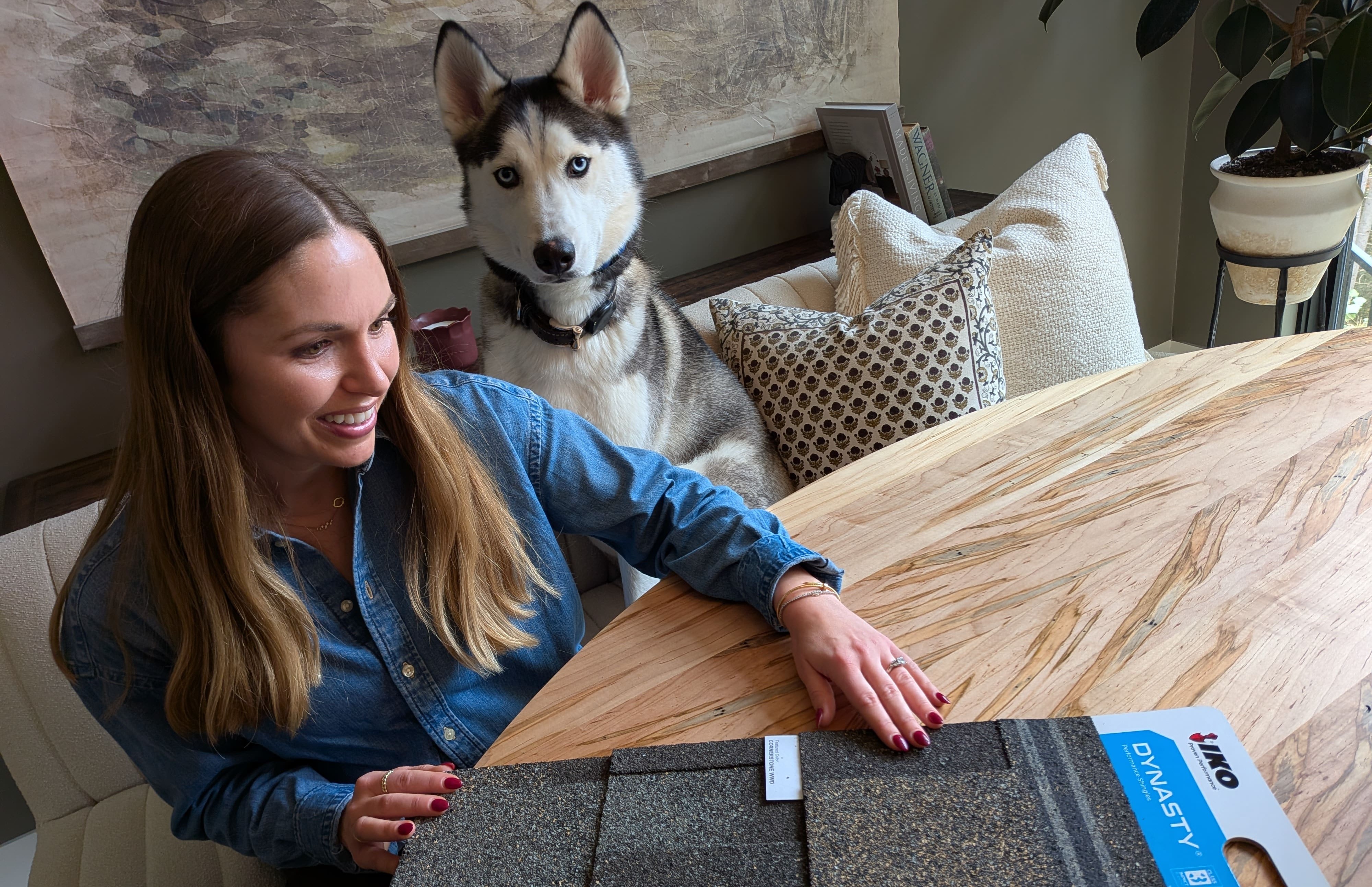 Mujer sonriente en una mesa de madera con un husky, mirando una muestra de teja IKO Dynasty