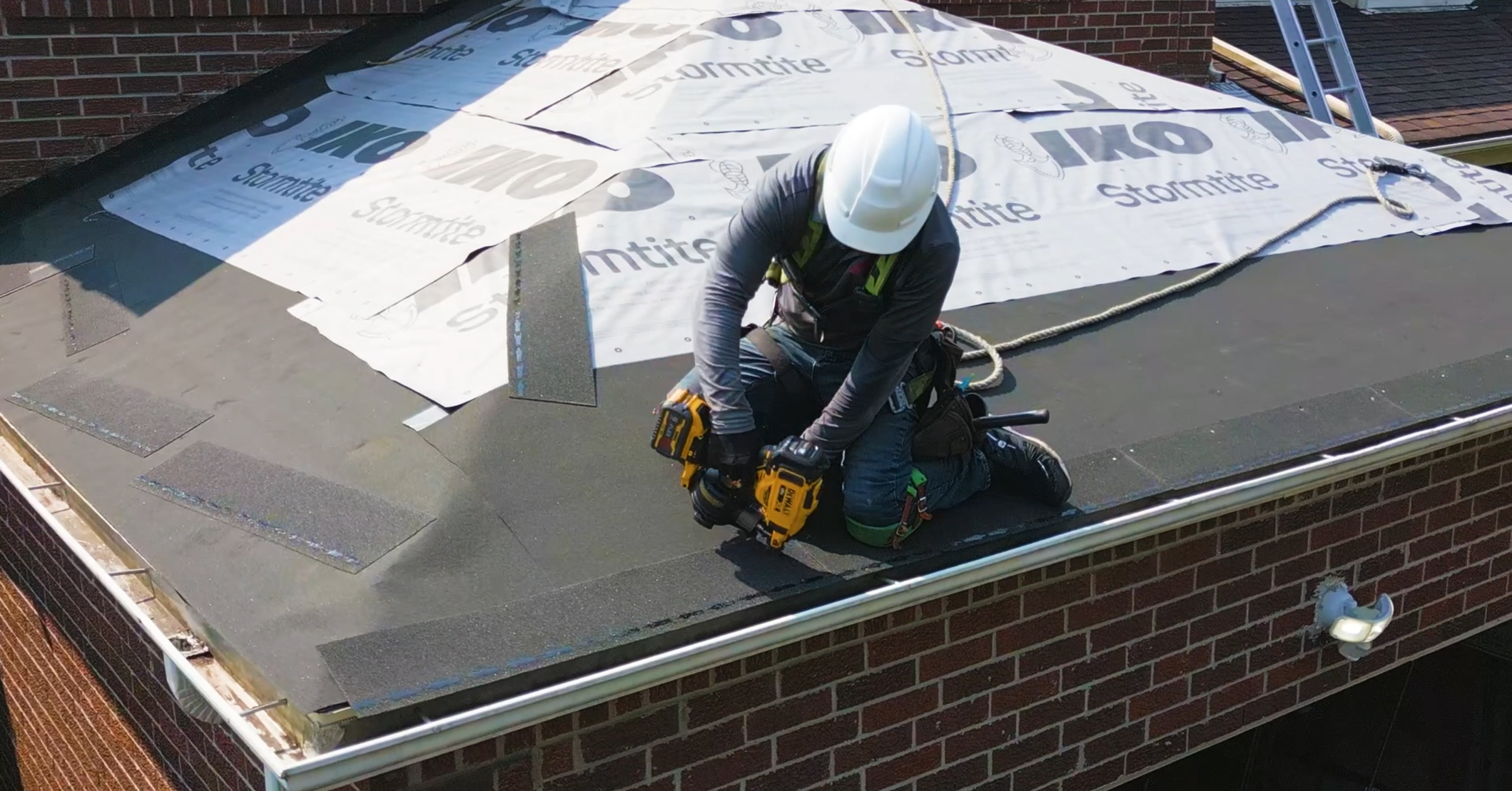 Alt text: Roofer installing shingles over black base layer with dual nail guns and IKO Stormtite visible on roof deck.