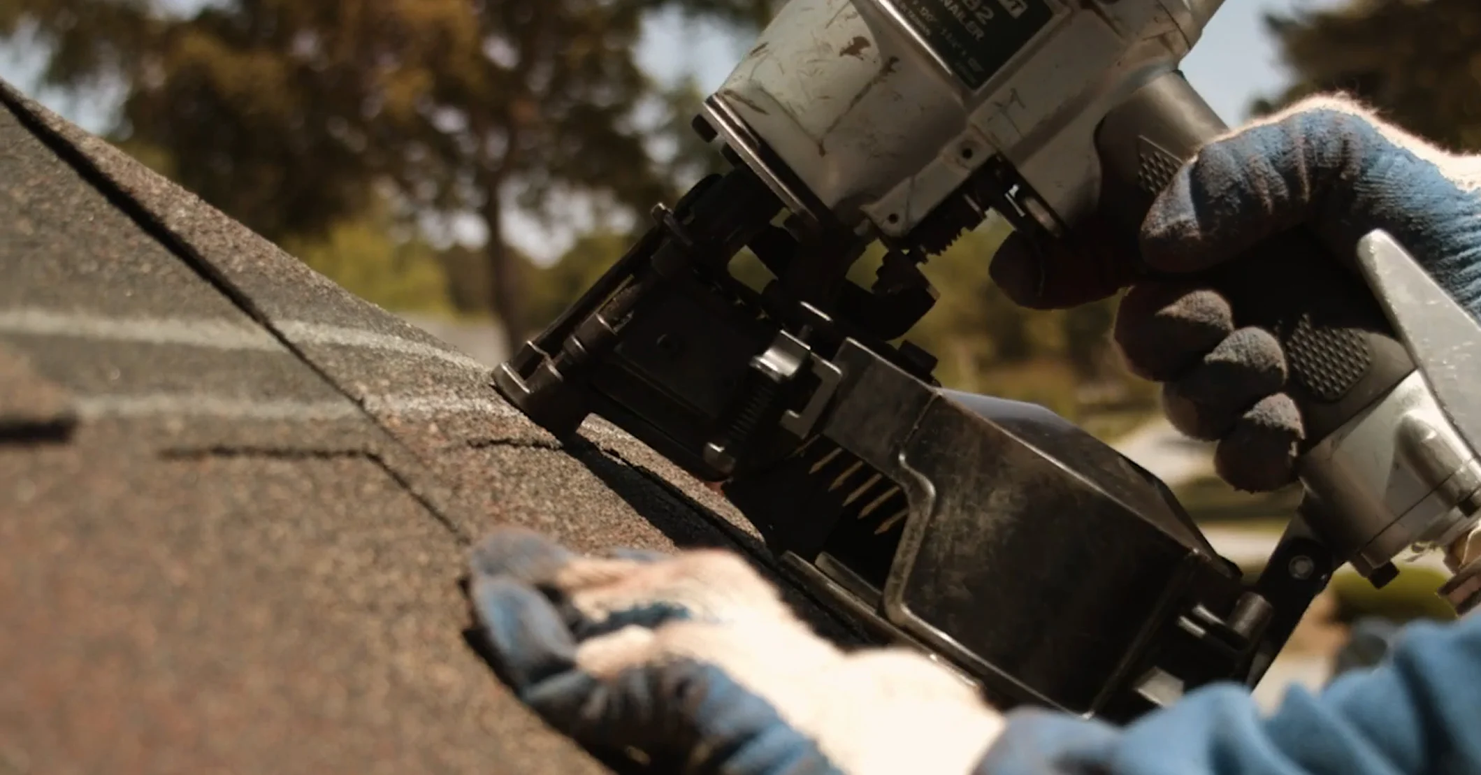 A roofer using a nail gun to fasten an asphalt shingle.