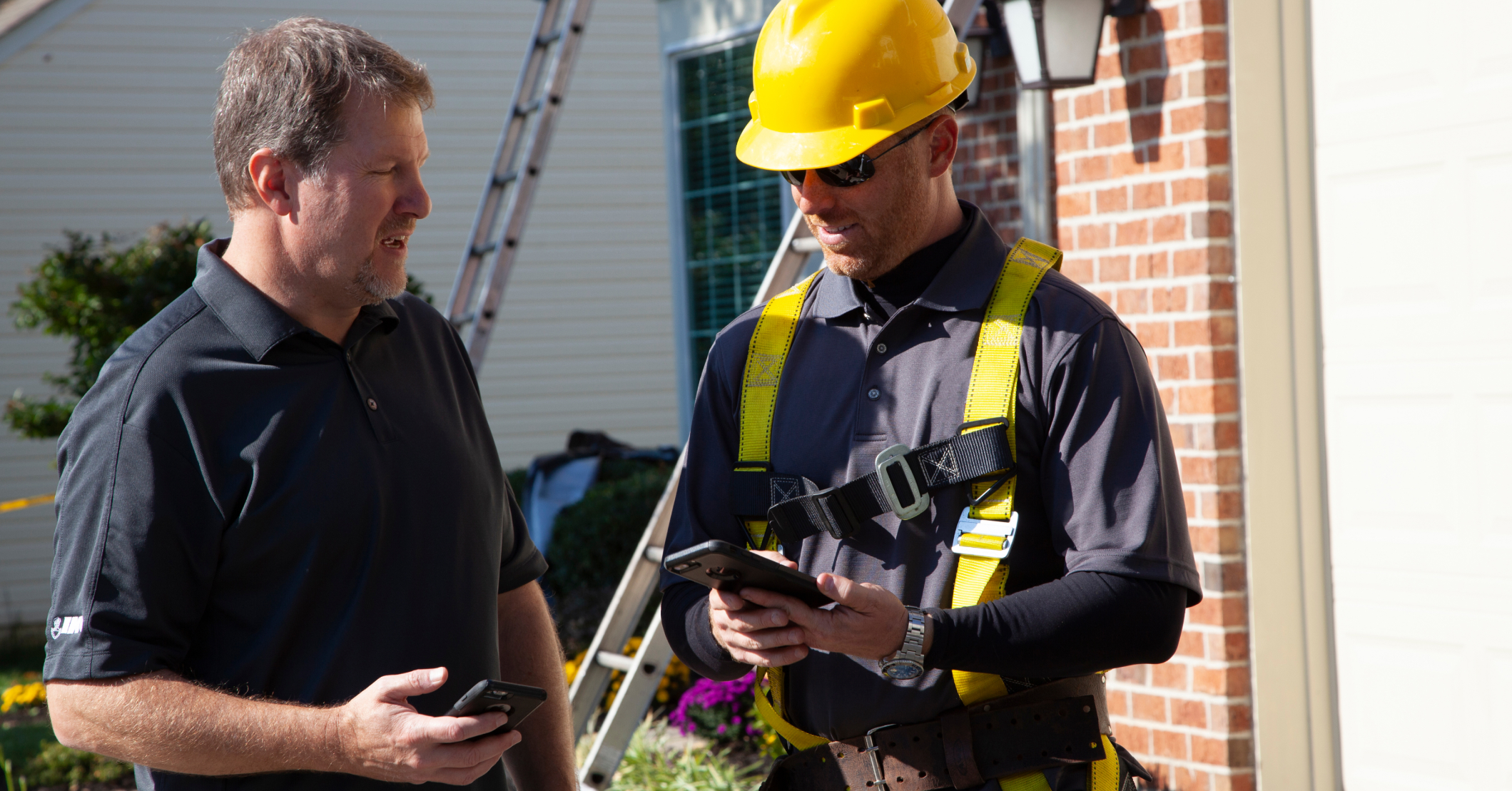 Homeowner consulting with a roofing contractor about a roof replacement claim. The contractor, wearing a hard hat and safety harness, reviews details on a smartphone.