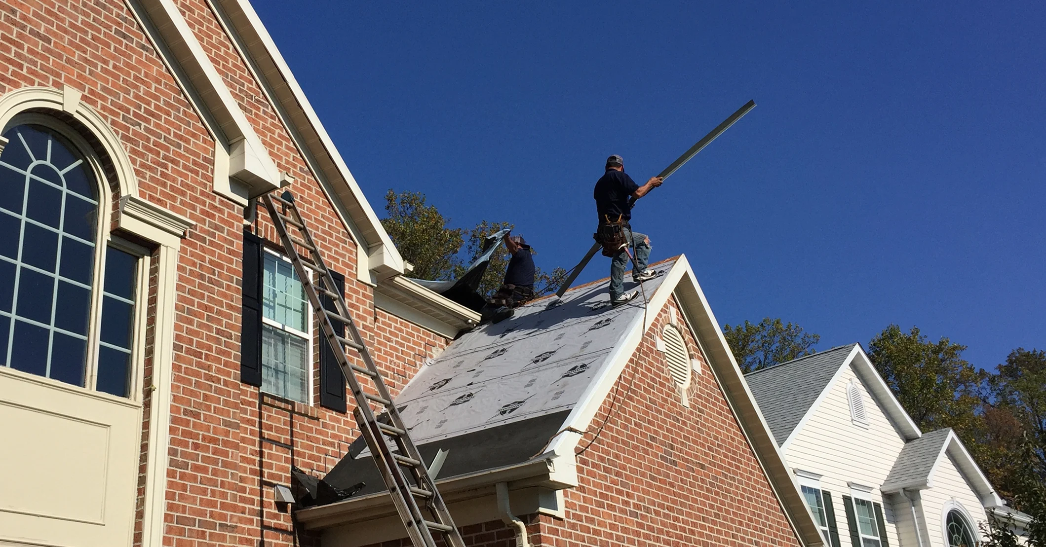Roofing contractors installing a new roof on a brick house using underlayment and metal flashing. Workers are wearing safety harnesses while preparing for shingle installation.