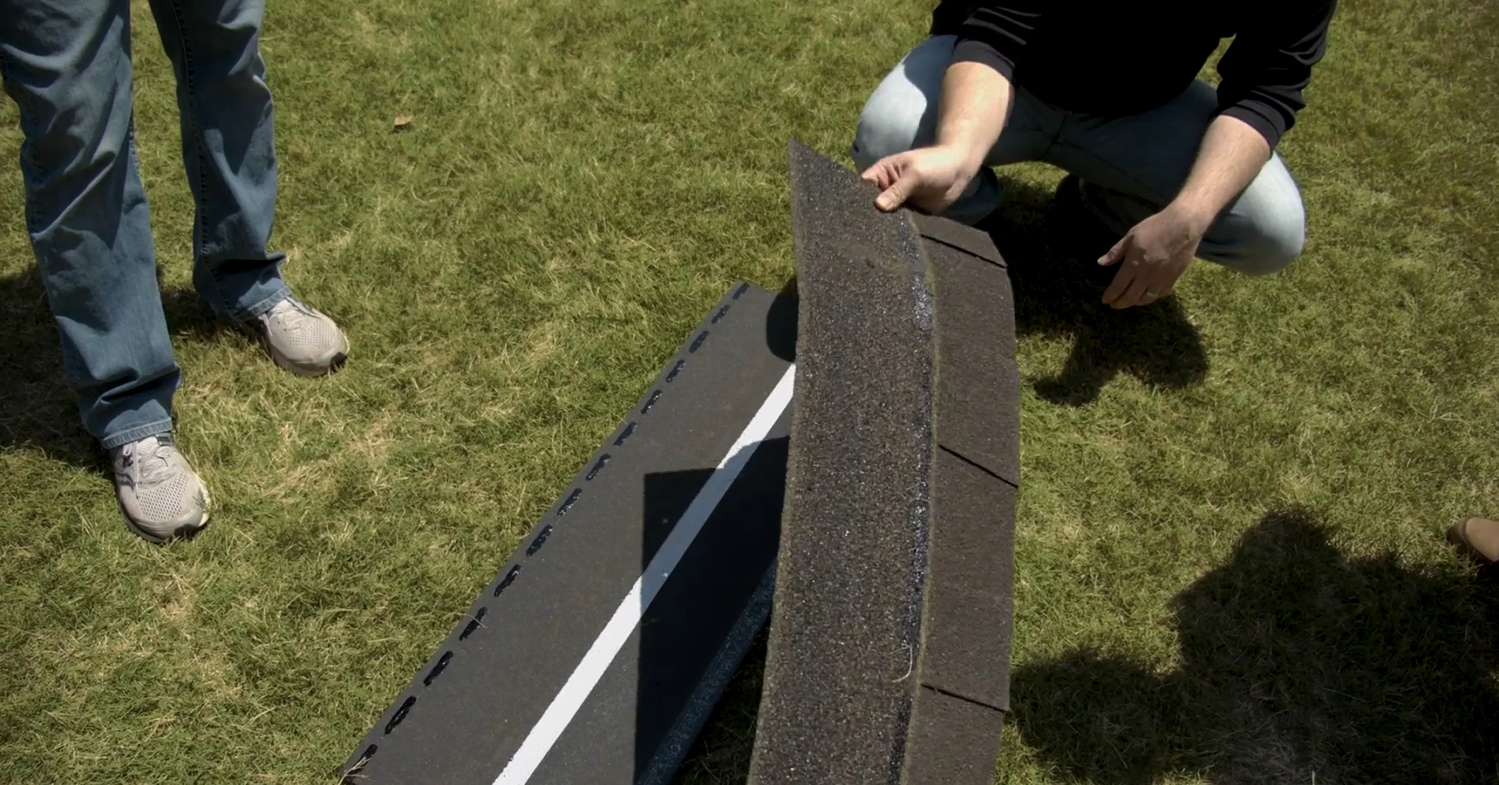 A close-up, high-angle shot of a person kneeling on grass while holding a dark-colored architectural shingle, demonstrating its features over a sample roof deck.