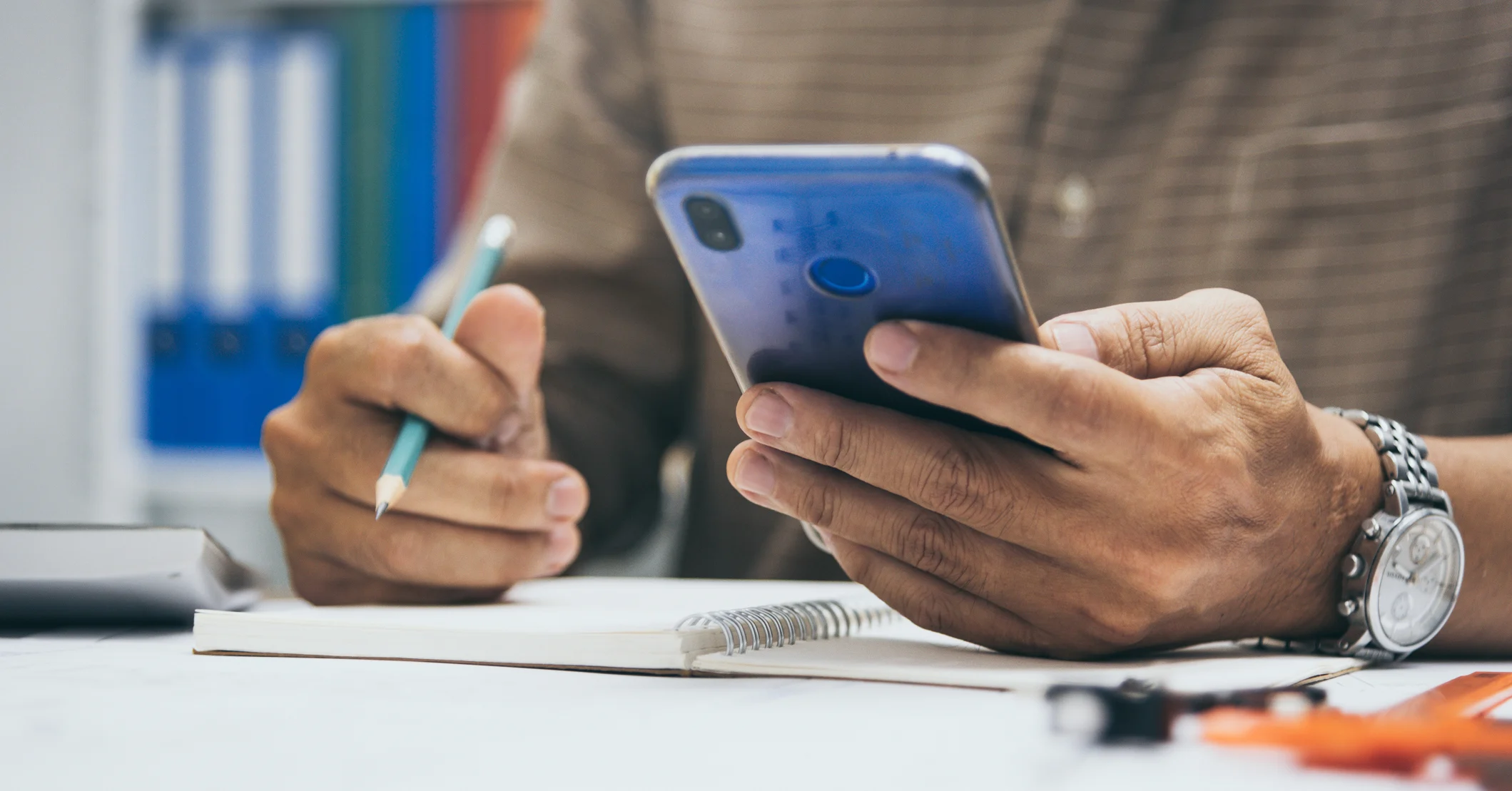 A business owner sits at a desk, holding a phone and a pen while looking at a notepad, appearing to be tracking business activities.