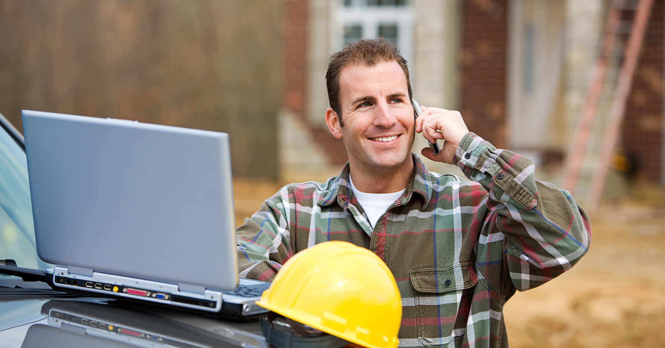 A smiling contractor in a plaid shirt talks on the phone while standing next to a laptop and a yellow hard hat at a job site.