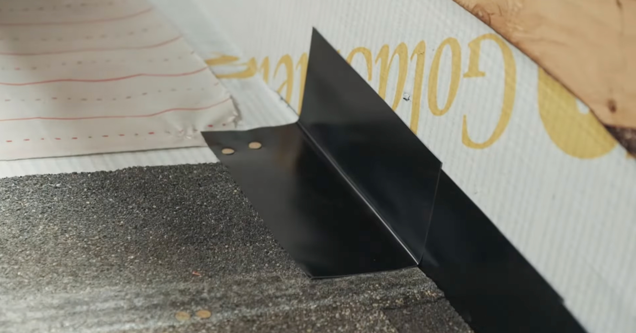 A close-up view of a black metal step flashing installed where dark asphalt shingles on a roof meet the gold-colored sheathing of a vertical wall.