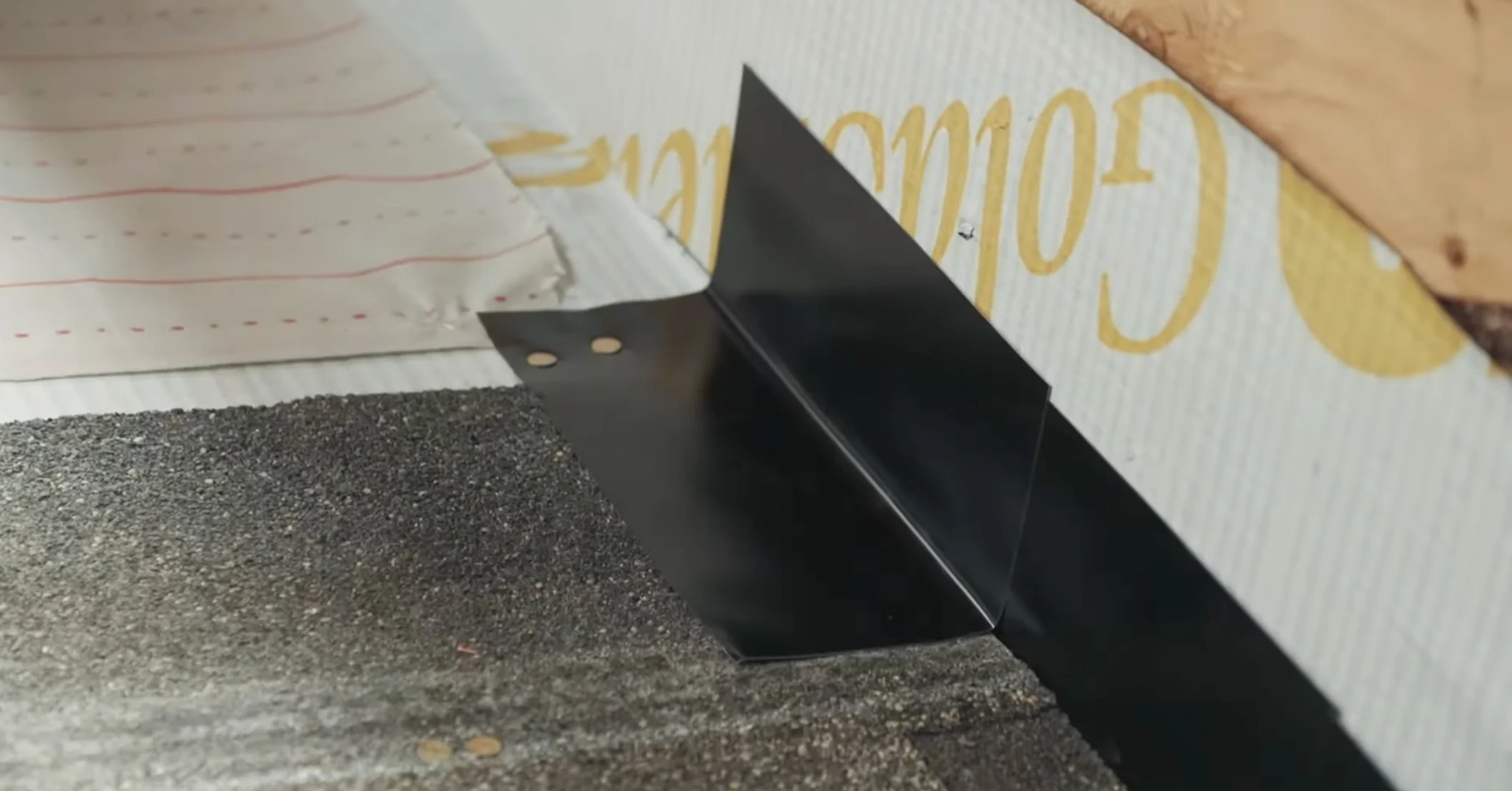 A close-up view of a black metal step flashing installed where dark asphalt shingles on a roof meet the gold-colored sheathing of a vertical wall.