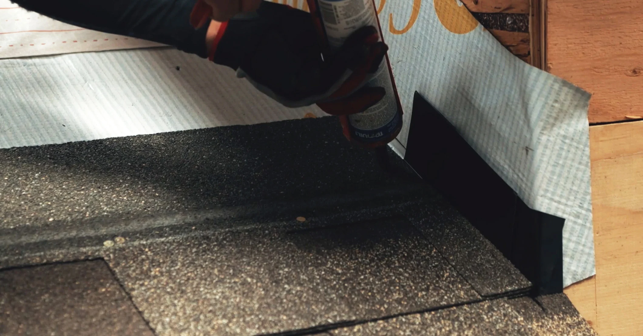 A close-up shot of a caulking gun applying a bead of sealant along the top edge of a black metal roof flashing where it meets the wall sheathing.