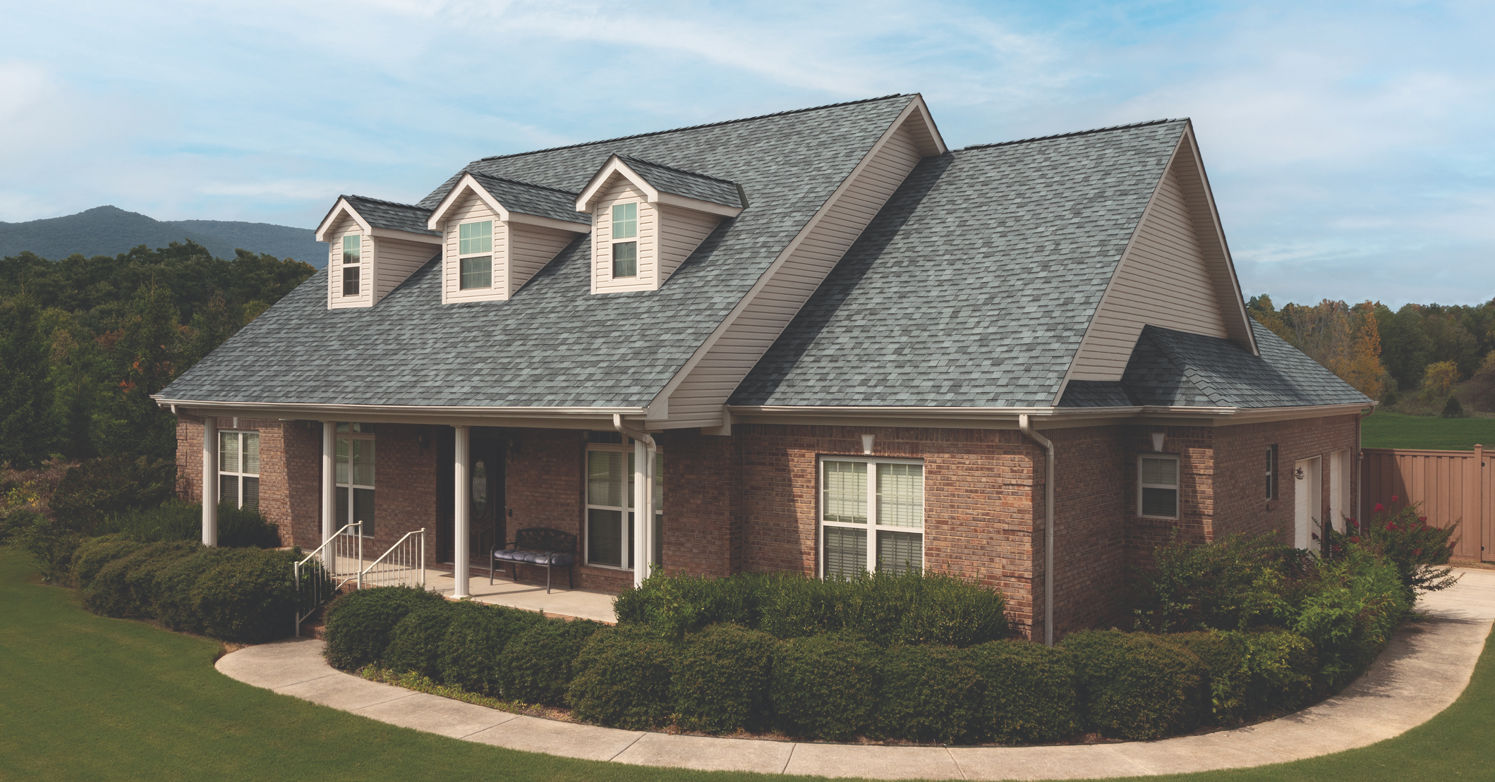 Red brick house with light siding accents, grey shingle roof, and three front-facing dormer windows. The home features white trim, a small front porch with seating, manicured hedges, and a curved concrete walkway.
