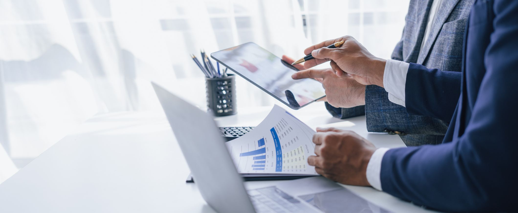 Two businessmen in suits review financial charts on a laptop and tablet.