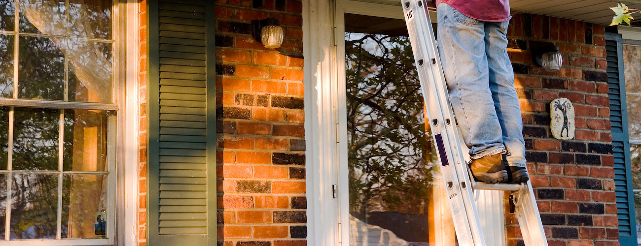 Person's legs standing on an aluminum ladder leaning against a brick house with windows and green shutters