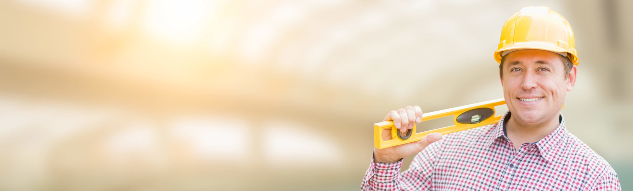 Smiling contractor in a yellow hard hat and plaid shirt holding a yellow level on his shoulder