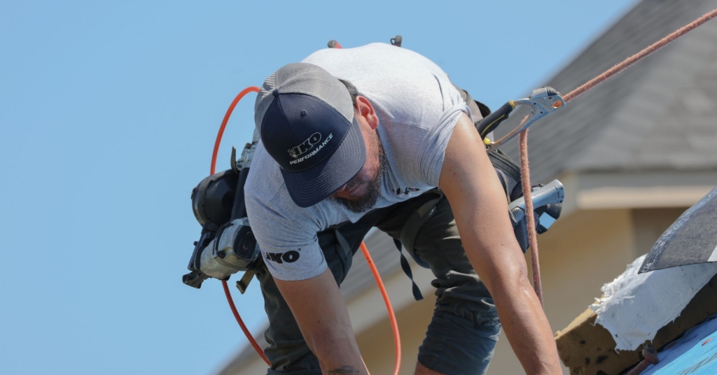 Un techador que est&aacute; utilizando ropa de IKO est&aacute; instalando tejas en un techo mientras lleva puesto un arn&eacute;s para techadores.
