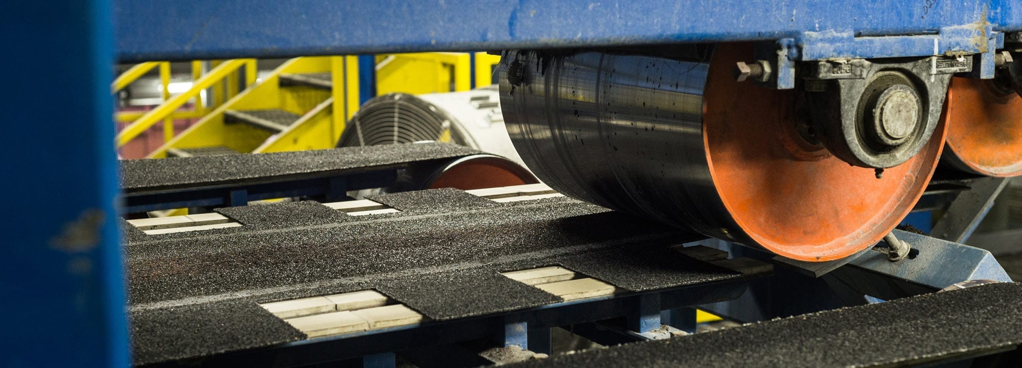 Asphalt shingles moving along a manufacturing line beneath large rollers.