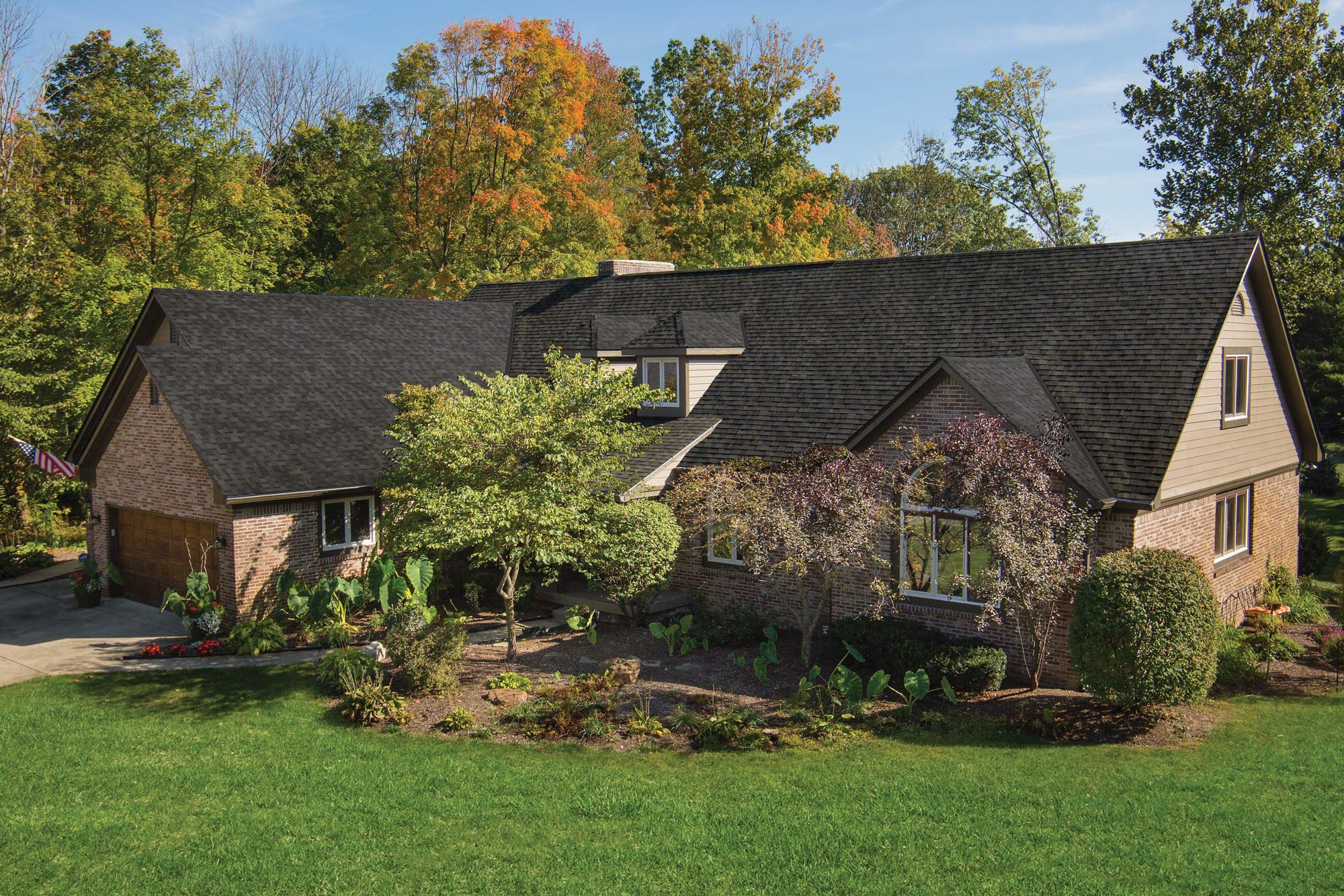 Brick and siding ranch home with a dark Dynasty Glacier shingle roof, landscaped garden and autumn trees