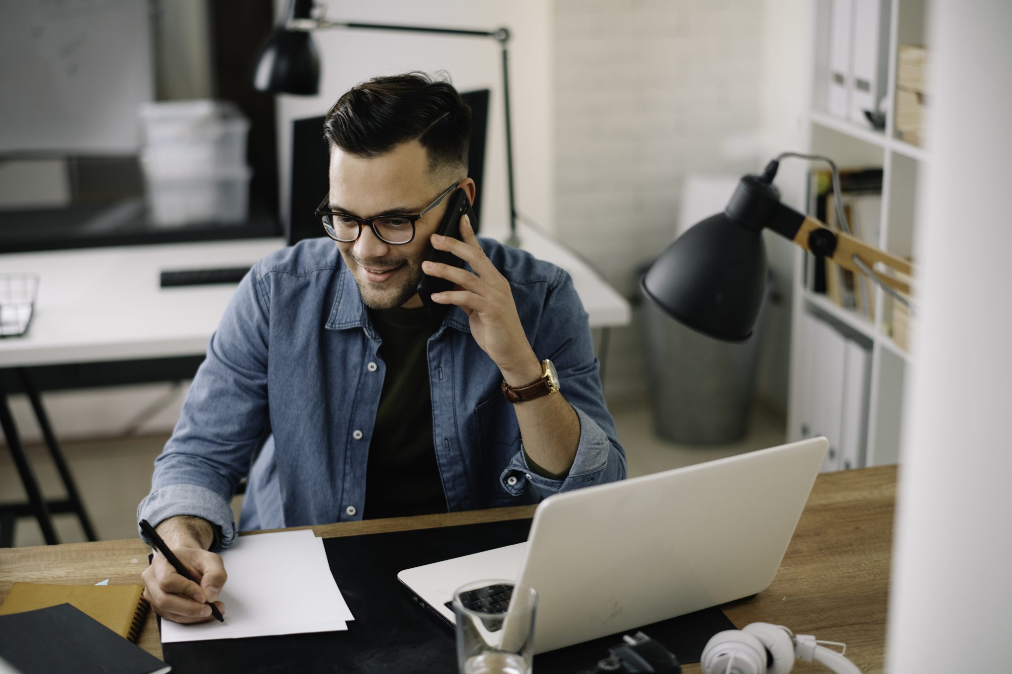 Smiling man in glasses talks on the phone and writes notes at a desk with a laptop.