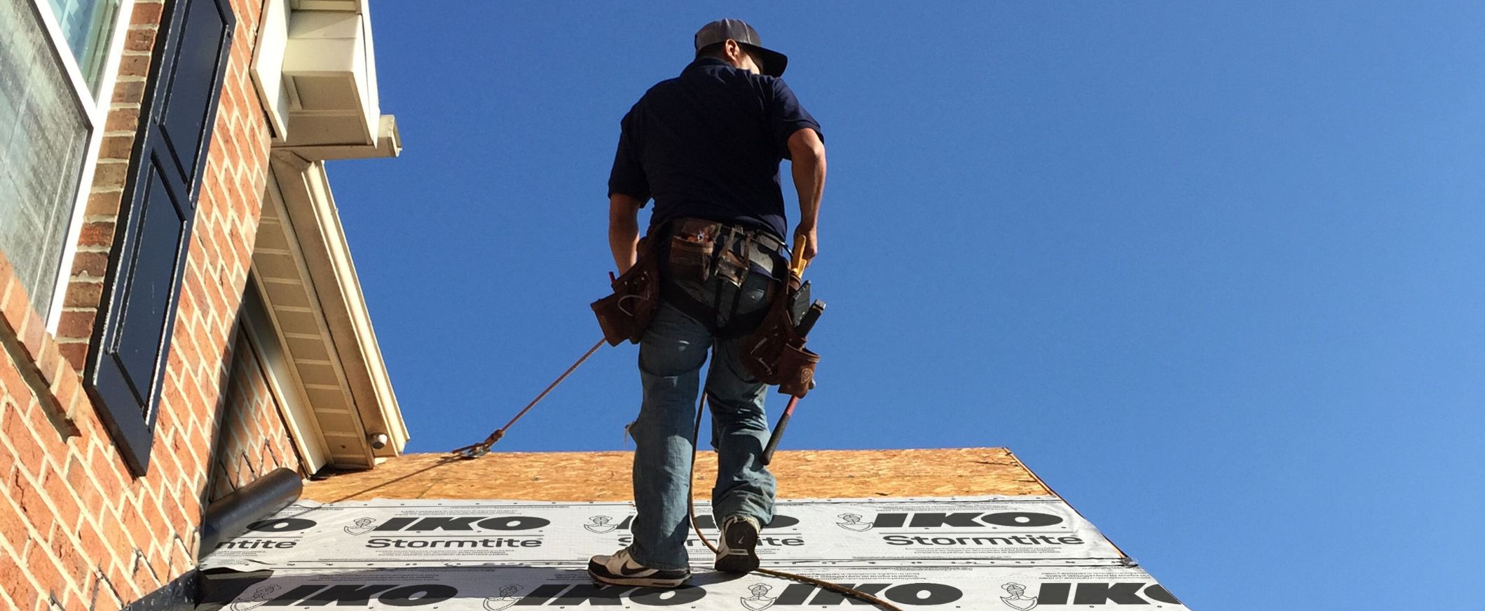 Professional roofer installing IKO Stormtite underlayment on a residential roof against a clear blue sky