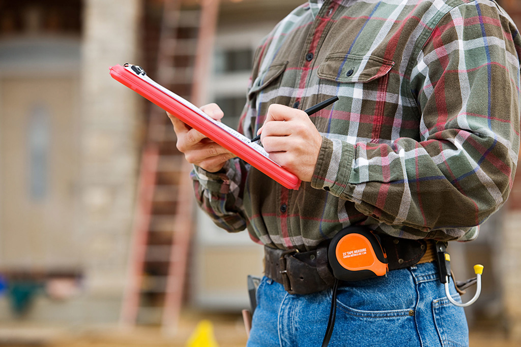 Inspector in a plaid shirt writes on a red clipboard with a tape measure on his belt