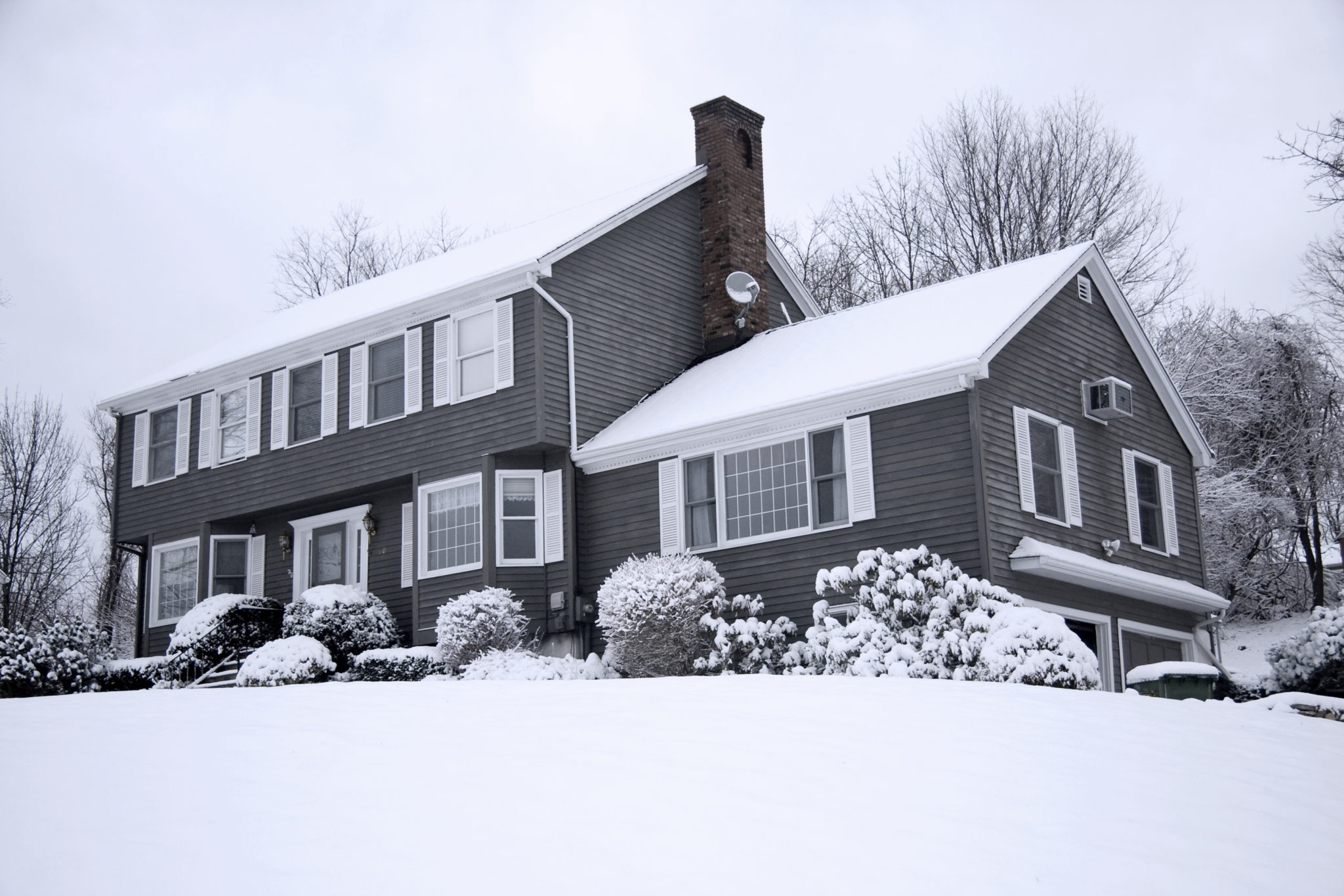 Two-story house with gray siding and a snow-covered roof, surrounded by snowy shrubs and bare trees