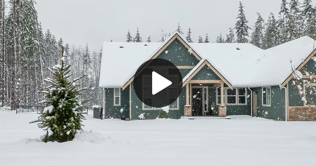 Home with a snow-covered roof surrounded by snowy evergreens, with a video play button