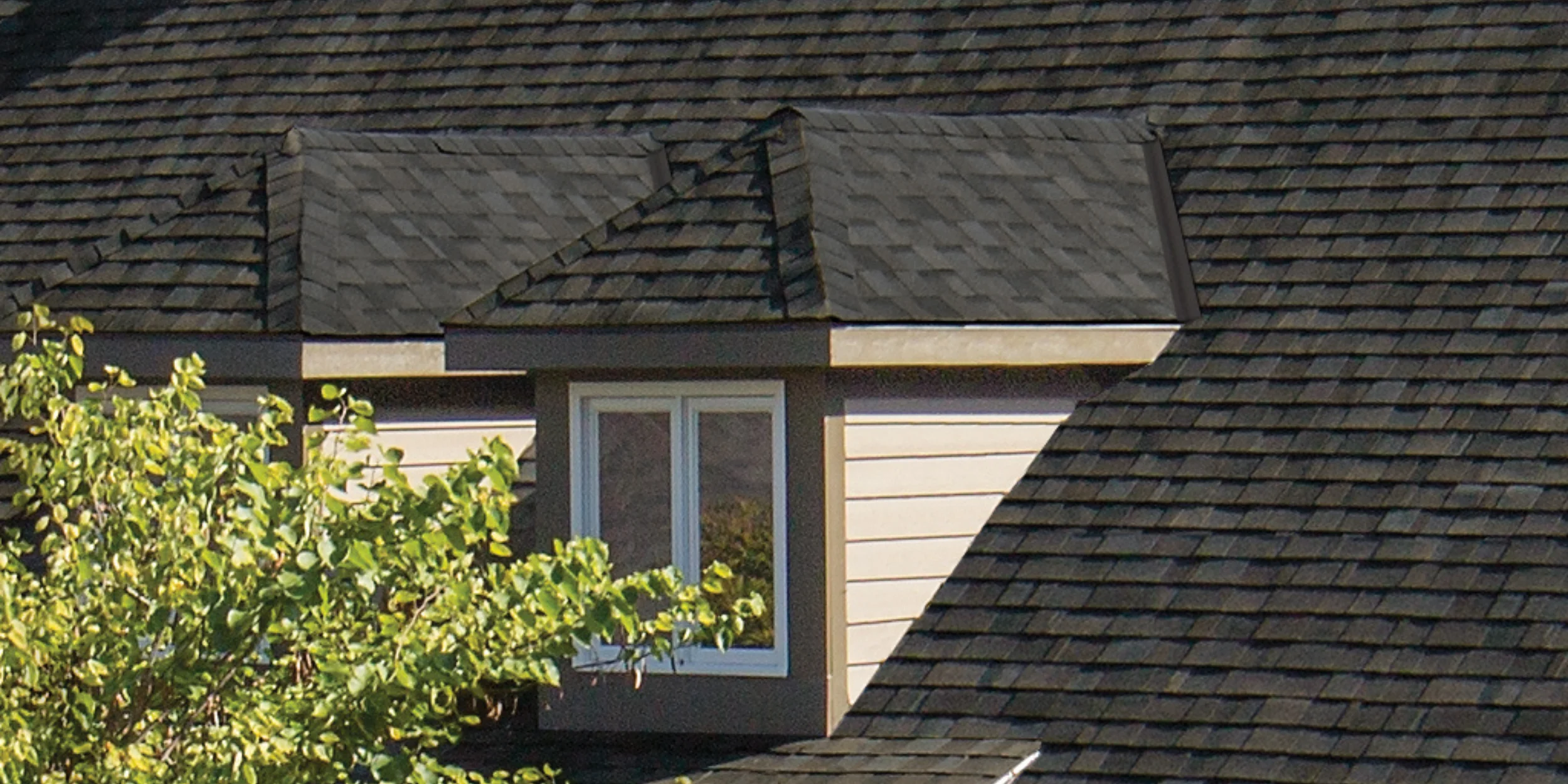 A hipped dormer with a three-sided sloped roof, blending into a dark shingle rooftop, housing a modern rectangular window.