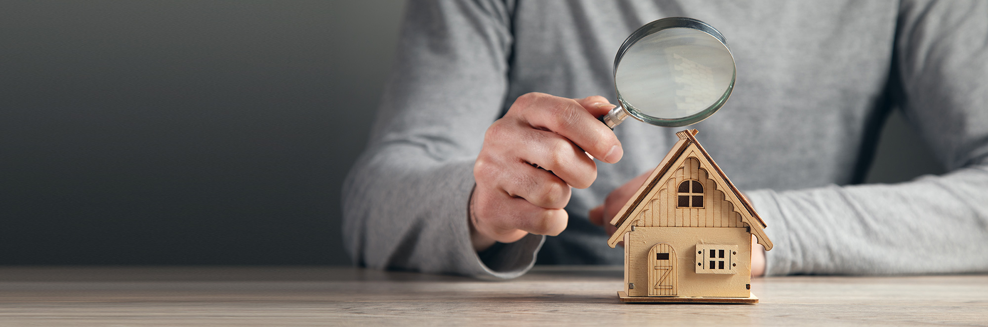 Hand holding a magnifying glass over a small wooden model house on a table.