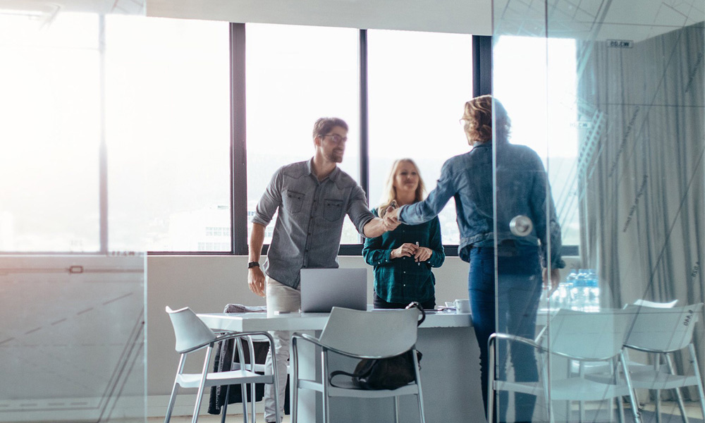 Two men and a woman shaking hands around a table with a laptop in a bright glass-walled office