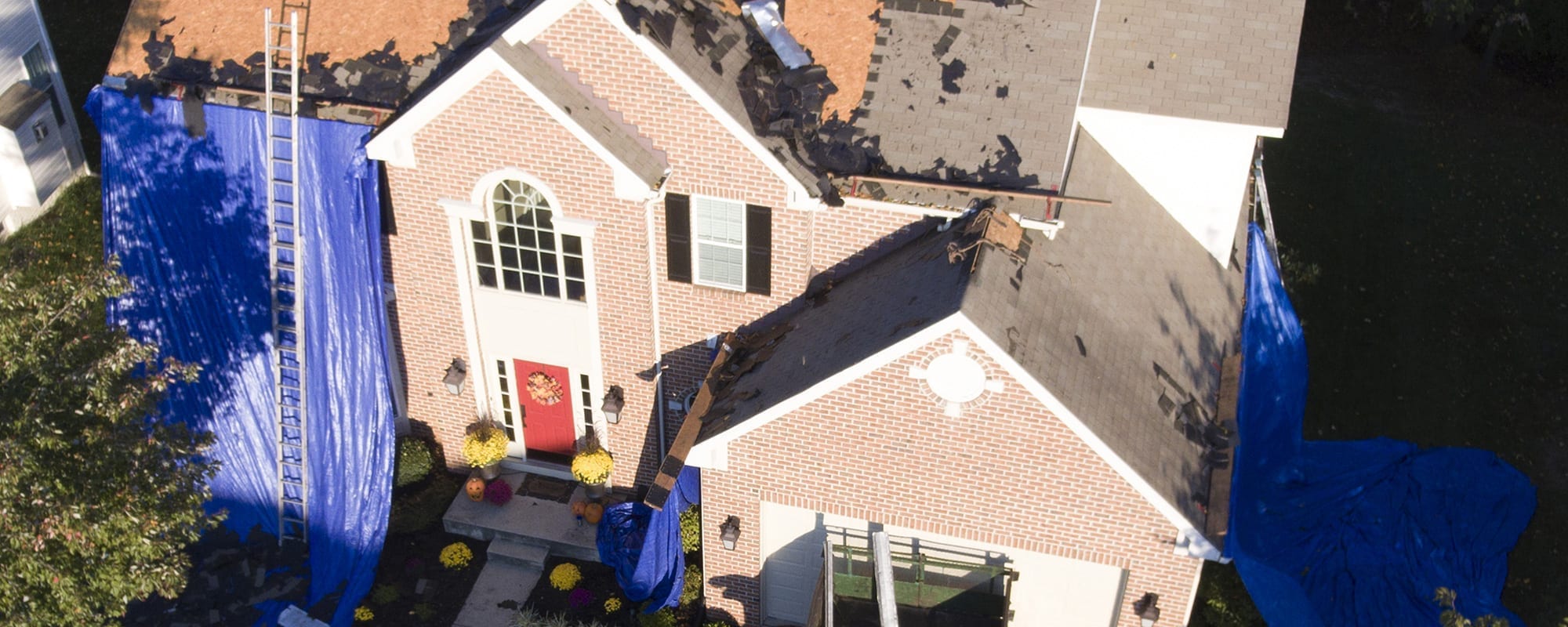 Aerial view of a brick house with a partly stripped roof, blue tarps and a ladder
