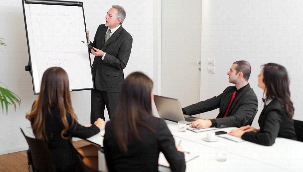 Man in a suit presents a graph on a whiteboard to three colleagues seated at a table