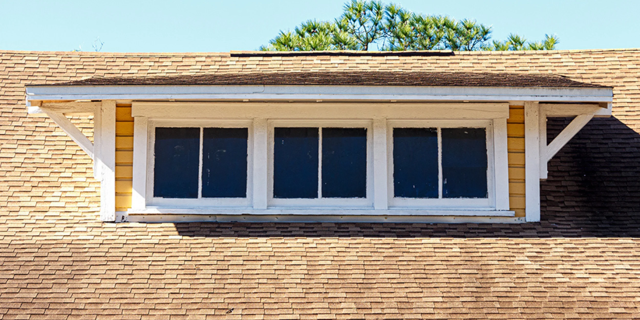 A shed dormer with a flat, single-slope roof and three vertical windows, extending from a gabled roof and supported by white brackets.