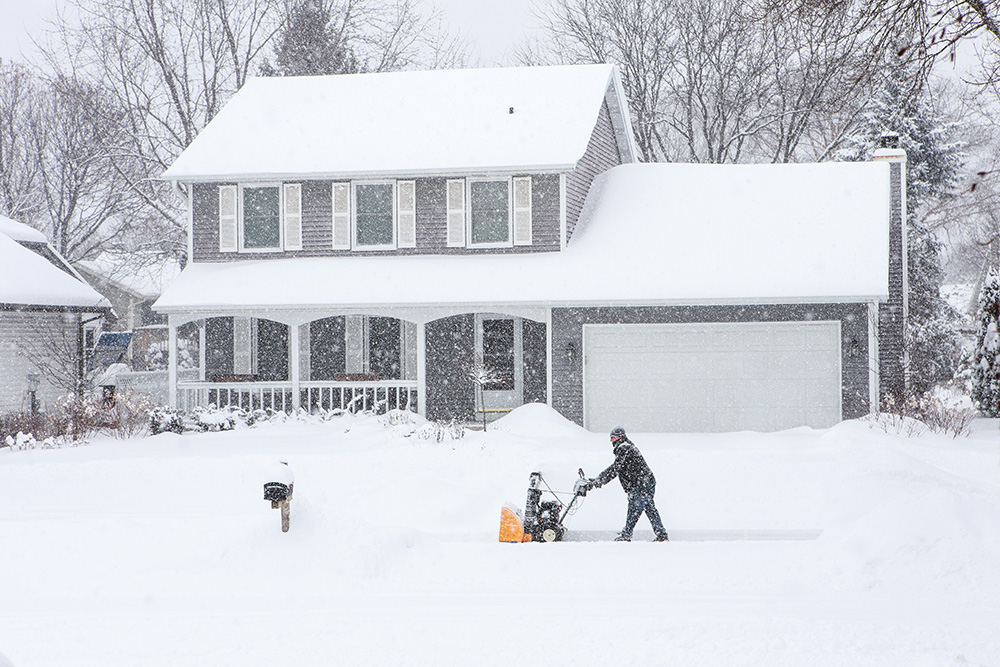 Person using a snow blower to clear a driveway in front of a snow-covered two-story house