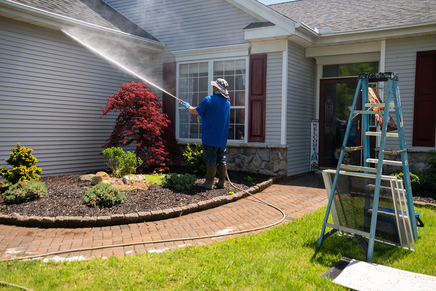 Man power washing the siding of a house, with a ladder and landscaped garden in front.